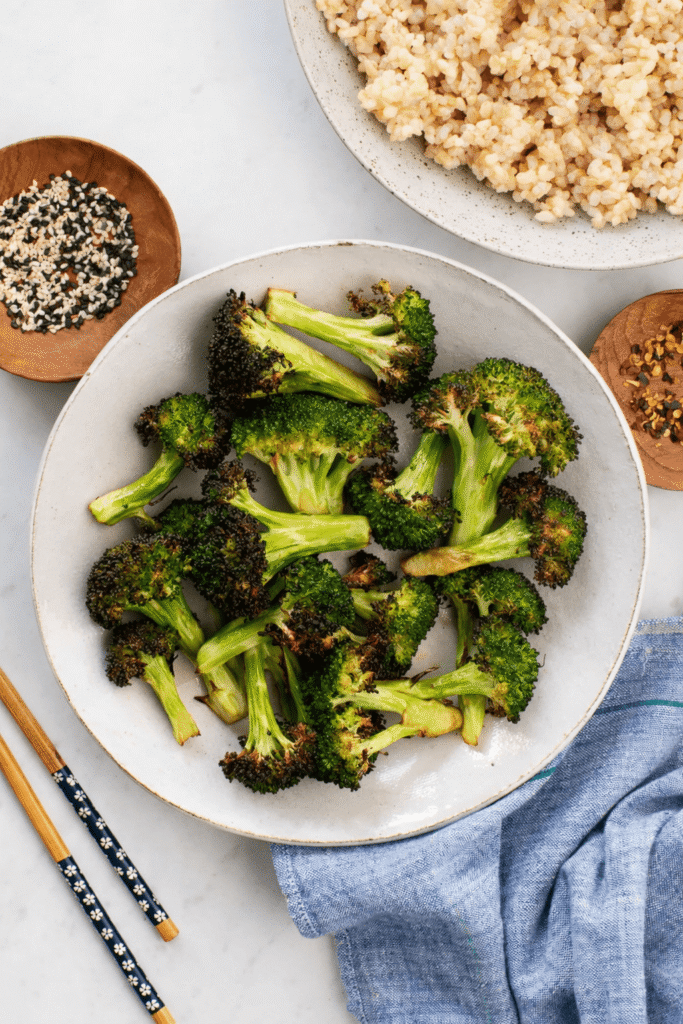 Gerösteter Brokkoli auf weißem Teller mit Vollkornreis, Sesam und Chiliflocken auf hellem Untergrund