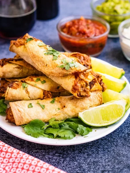 Eine Nahaufnahme von einem Teller mit knusprigen Hähnchen-Taquitos, garniert mit Koriander und Limettenstücken, serviert mit Salsa, Guacamole und Sauerrahm im Hintergrund.