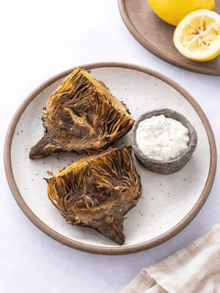 „Knusprig geröstete Air Fryer Artischockenhälften auf einem rustikalen Teller, serviert mit einem Kräuterdip und einer Zitrone im Hintergrund.“