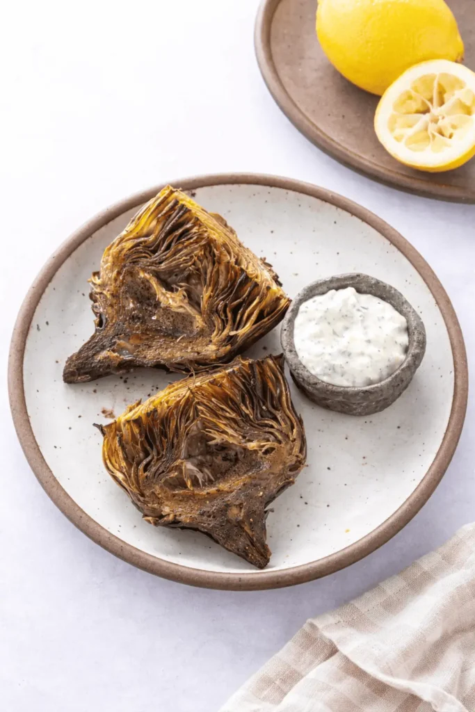 „Knusprig geröstete Air Fryer Artischockenhälften auf einem rustikalen Teller, serviert mit einem Kräuterdip und einer Zitrone im Hintergrund.“