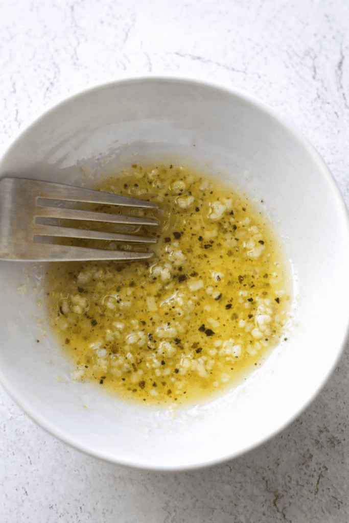 Geschmolzene Knoblauch-Zitronen-Butter mit schwarzem Pfeffer in einer weißen Schüssel, angerührt mit einer Gabel.